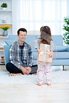 Cute little girl, daughter, sister gives a gift box to young dad father or brother. Both are smiling. Father`s day holiday concep