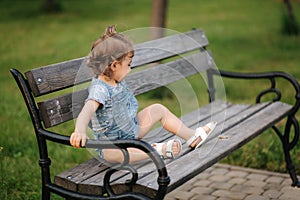 Cute little girl in denim climbs on the bench in the park. Happy smiled kid on the bench