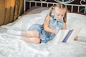 Cute little girl in denim sundress reading book on bed at home