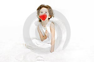 A cute little girl in dress and white wings, sitting on a fur, holding a red heart on her lips, over white background.