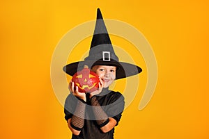 Cute little girl dressed in a witch costume holds a pumpkin jack lantern on a yellow background. Halloween celebration