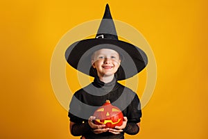 Cute little girl dressed in a witch costume holds a pumpkin jack lantern on a yellow background. Halloween celebration