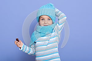 Cute little girl dresses striped casual shirt, warm hat with pom pom and scarf, keeping hand on her cap, holding candy in hansd,
