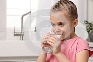 Cute little girl drinking fresh water from glass in kitchen