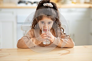 Cute Little Girl Drinking Water From Glass, Sitting At Table In Kitchen