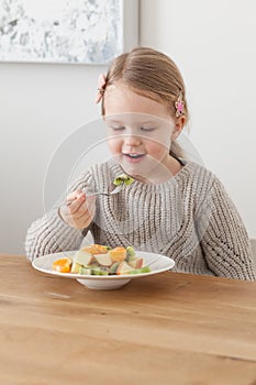 Cute little girl eating a fruit salad in design dining room. Child having a dinner at home. Healthy nutrition for small
