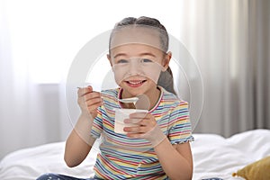 Cute little girl eating tasty yogurt on bed