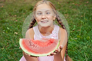 Cute little girl eating watermelon on the grass in summer time. with ponytail long hair and toothy smile sitting on grass and enjo