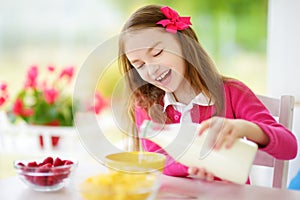 Cute little girl enjoying her breakfast at home. Pretty child eating corn flakes and raspberries and drinking milk before school.