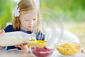Cute little girl enjoying her breakfast at home. Pretty child eating corn flakes and raspberries and drinking milk before school.