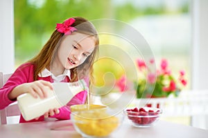 Cute little girl enjoying her breakfast at home. Pretty child eating corn flakes and raspberries and drinking milk before school.