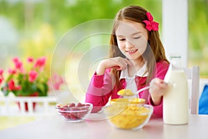 Cute little girl enjoying her breakfast at home. Pretty child eating corn flakes and raspberries and drinking milk before school.