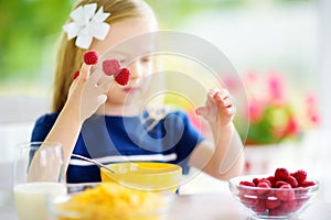 Cute little girl enjoying her breakfast at home. Pretty child eating corn flakes and raspberries and drinking milk before school.