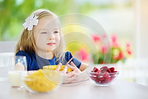 Cute little girl enjoying her breakfast at home. Pretty child eating corn flakes and raspberries and drinking milk before school.