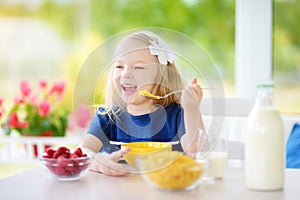 Cute little girl enjoying her breakfast at home. Pretty child eating corn flakes and raspberries and drinking milk before school.