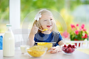 Cute little girl enjoying her breakfast at home. Pretty child eating corn flakes and raspberries and drinking milk before school.