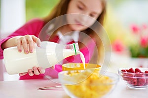 Cute little girl enjoying her breakfast at home. Pretty child eating corn flakes and raspberries and drinking milk before school.