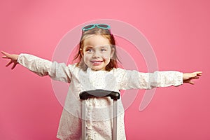 Cute little girl extended arms, depicts a plane, smiling happily, stands near suitcase on pink.