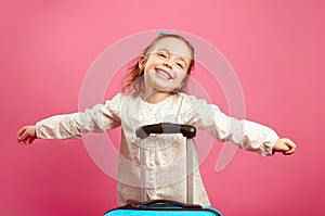 Cute little girl extended arms, depicts a plane, smiling happily, stands near suitcase on pink.