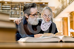 Cute little girl in eyeglasses sitting at the table in ancient library, hugging her grandfather and reading book