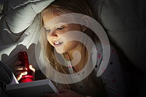 Cute little girl with flashlight reading book in bed under blanket