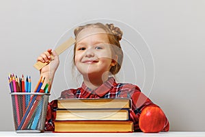 A cute little girl with glasses is sitting at the table, holding a ruler in her hand. Schoolgirl is surrounded by books.