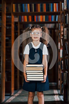 Cute little girl in glasses stands in the library full of books. Conception of education