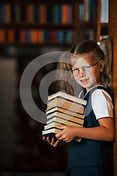 Cute little girl in glasses stands in the library full of books. Conception of education