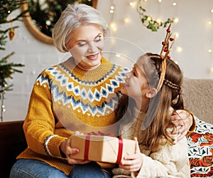 Little girl granddaughter giving Christmas gift box to smiling grandmother during winter holidays