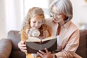 Cute little girl granddaughter reading book with positive senior grandmother