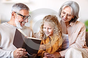 Cute little girl granddaughter smiling when reading book with senior grandparents