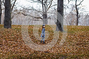 A cute little girl in grey coat and gray hat near the thick old oak in the oak wood at the sunset