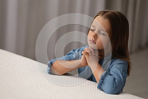 Cute little girl with hands clasped together saying bedtime prayer in bedroom. Space for text