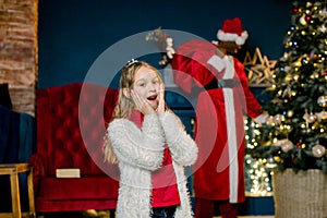Cute little girl is happy and excited standing in cozy decorated room. African Santa Claus came and puts presents at the