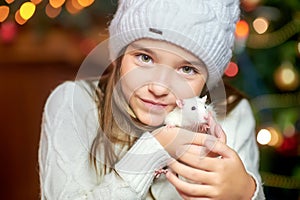 Cute little girl in a hat holds in her arms and hugs a funny white rat on a background of bright holiday lights.