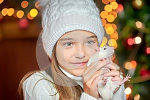 Cute little girl in a hat holds in her arms and hugs a funny white rat on a background of bright holiday lights