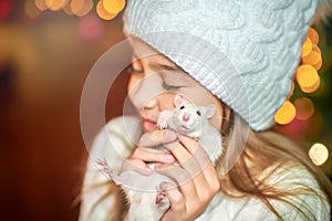 Cute little girl in a hat holds in her arms and hugs a funny white rat on a background of bright holiday lights