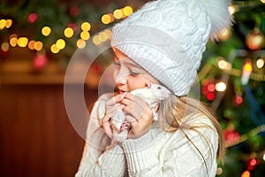 Cute little girl in a hat holds in her arms and hugs a funny white rat on a background of bright holiday lights