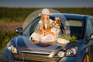 Cute little girl in a hat sitting on the hood of the car next to a dog and a bouquet of flowers. Summer vacation.