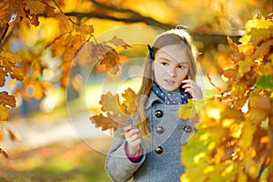 Cute little girl having fun on beautiful autumn day. Happy child playing in autumn park. Kid gathering yellow fall foliage.