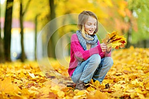 Cute little girl having fun on beautiful autumn day. Happy child playing in autumn park. Kid gathering yellow fall foliage.