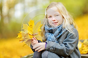 Cute little girl having fun on beautiful autumn day. Happy child playing in autumn park. Kid gathering yellow fall foliage.