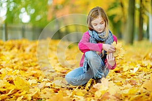 Cute little girl having fun on beautiful autumn day. Happy child playing in autumn park. Kid gathering yellow fall foliage.