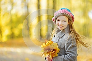 Cute little girl having fun on beautiful autumn day. Happy child playing in autumn park. Kid gathering yellow fall foliage.