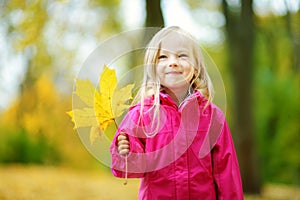 Cute little girl having fun on beautiful autumn day. Happy child playing in autumn park. Kid gathering yellow fall foliage.