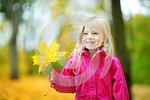 Cute little girl having fun on beautiful autumn day. Happy child playing in autumn park. Kid gathering yellow fall foliage.