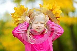 Cute little girl having fun on beautiful autumn day. Happy child playing in autumn park. Kid gathering yellow fall foliage.