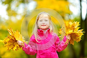 Cute little girl having fun on beautiful autumn day. Happy child playing in autumn park. Kid gathering yellow fall foliage.