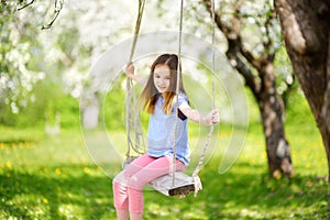 Cute little girl having fun on a swing in blossoming old apple tree garden outdoors on sunny spring day.