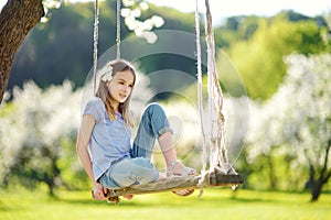 Cute little girl having fun on a swing in blossoming old apple tree garden outdoors on sunny spring day.